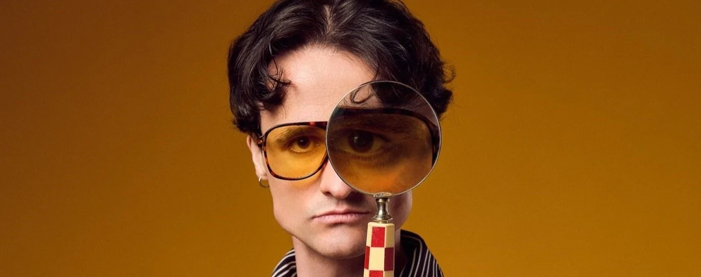 A man with dark wavy hair and wearing yellow-tinted sunglasses looks directly at the camera, holding a magnifying glass in front of their face revealing their eye.