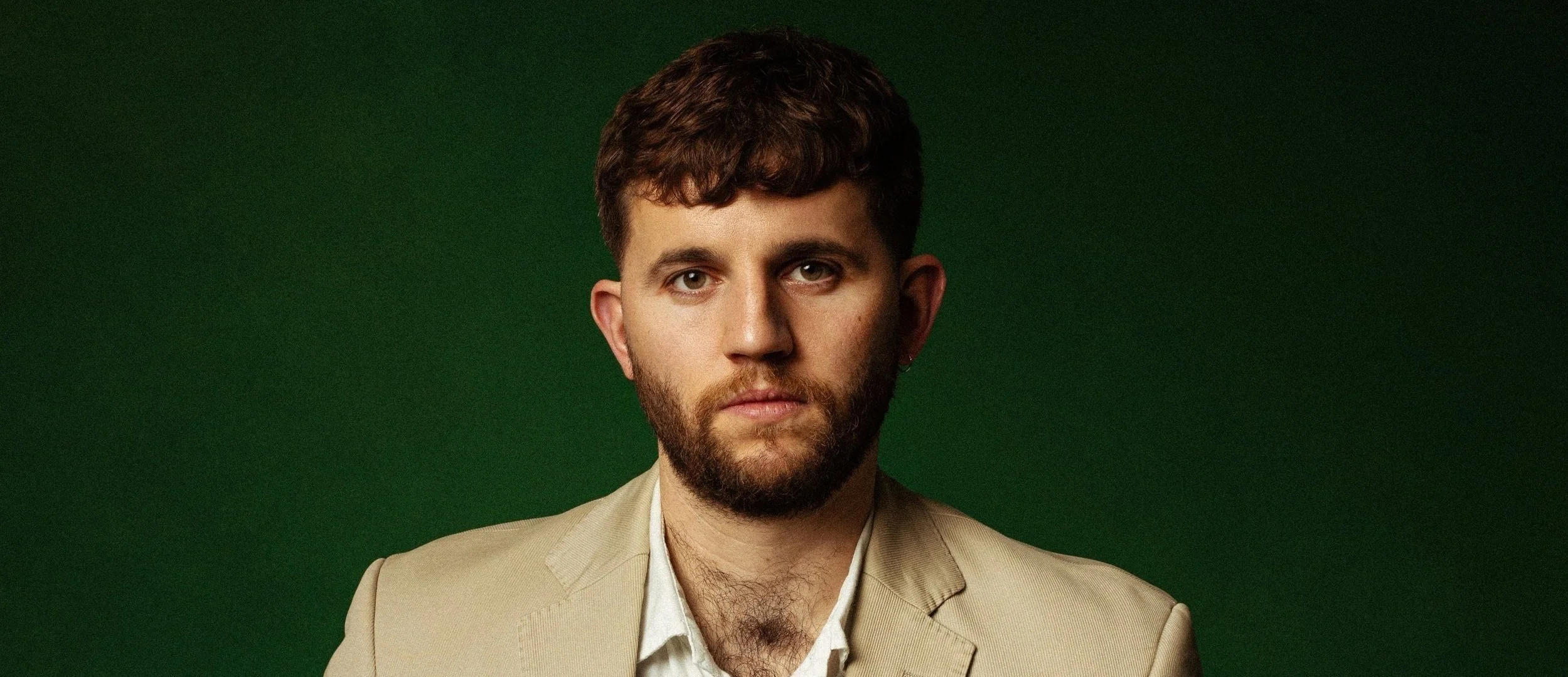 Portrait of a young man with short brown hair, a beard, wearing a beige blazer over a white shirt, against a dark green background.