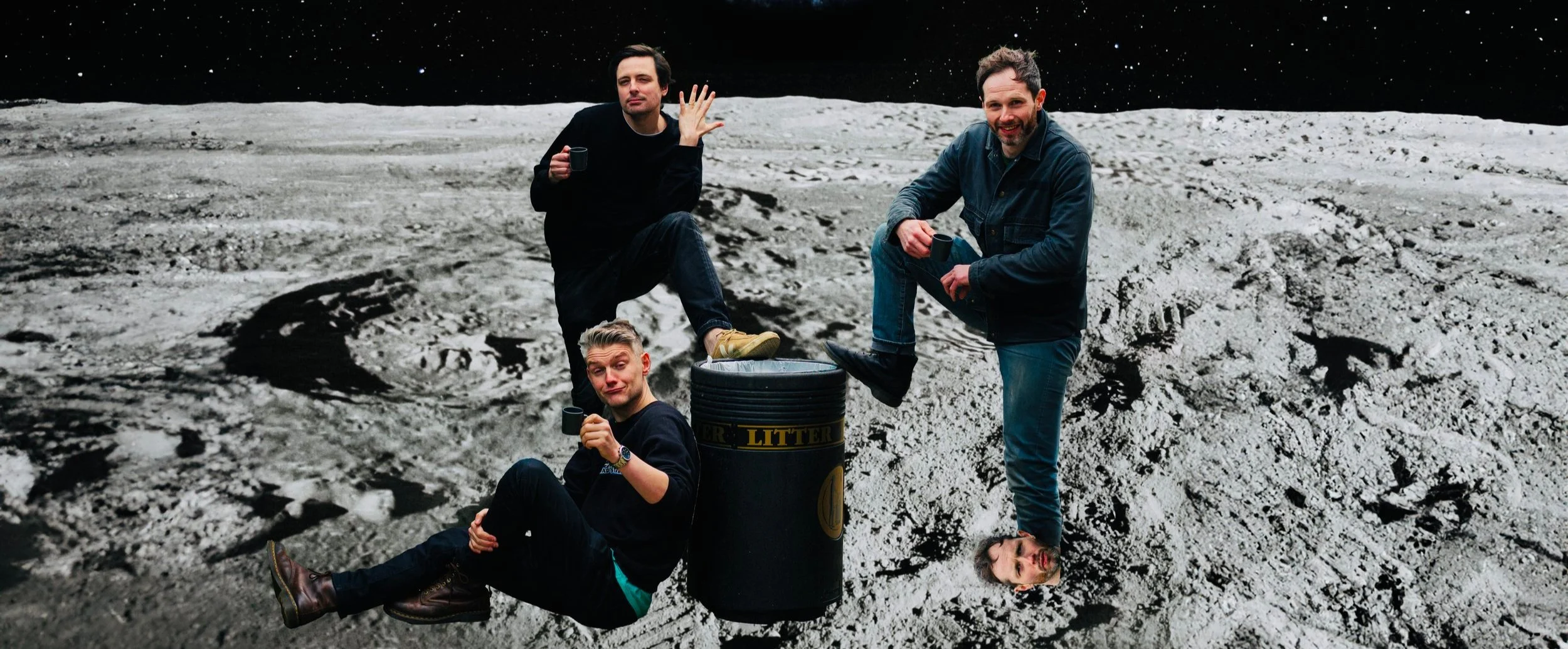 Three men on the moon's surface, with one men sitting and the other two standing, holding mugs. One of the standing men is smiling, and the sitting man appears relaxed. The moon's surface is gray and rocky, with Earth visible as a black, starry sky in the background.