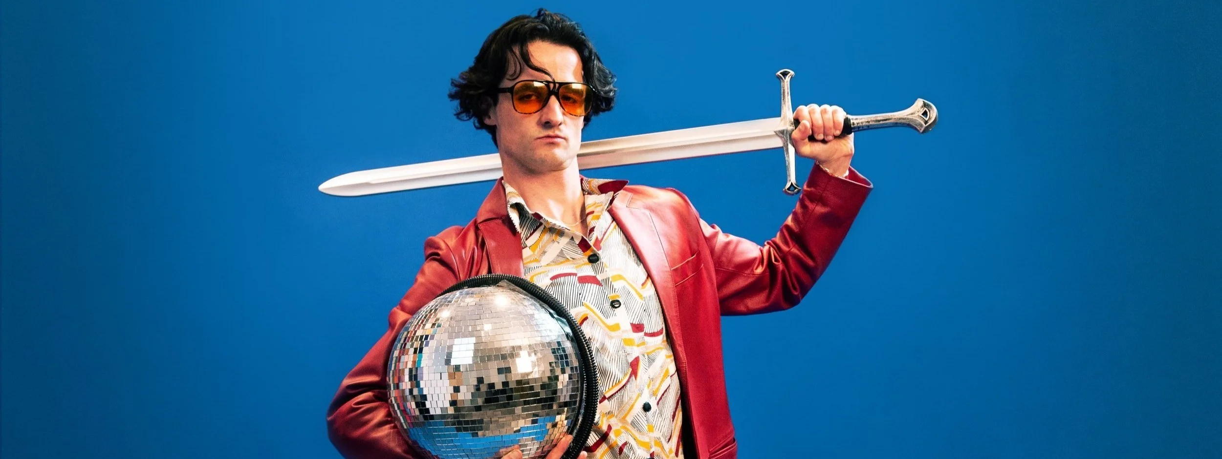 A man with curly dark hair wearing orange-tinted sunglasses, a red leather jacket, and a colorful patterned shirt, holding a large disco ball in one hand and a sword resting on his shoulder against a plain blue background.