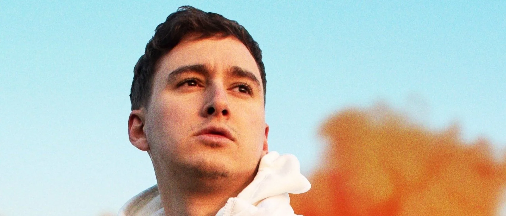 Close-up of a young man with short dark hair, looking into the distance with a thoughtful expression, against a blue sky background.