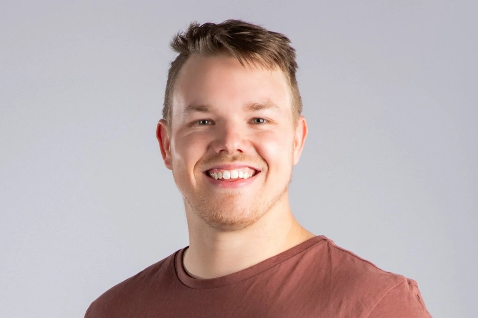 A young man with short brown hair, light skin, and a big smile, wearing a maroon T-shirt, standing against a light gray background.