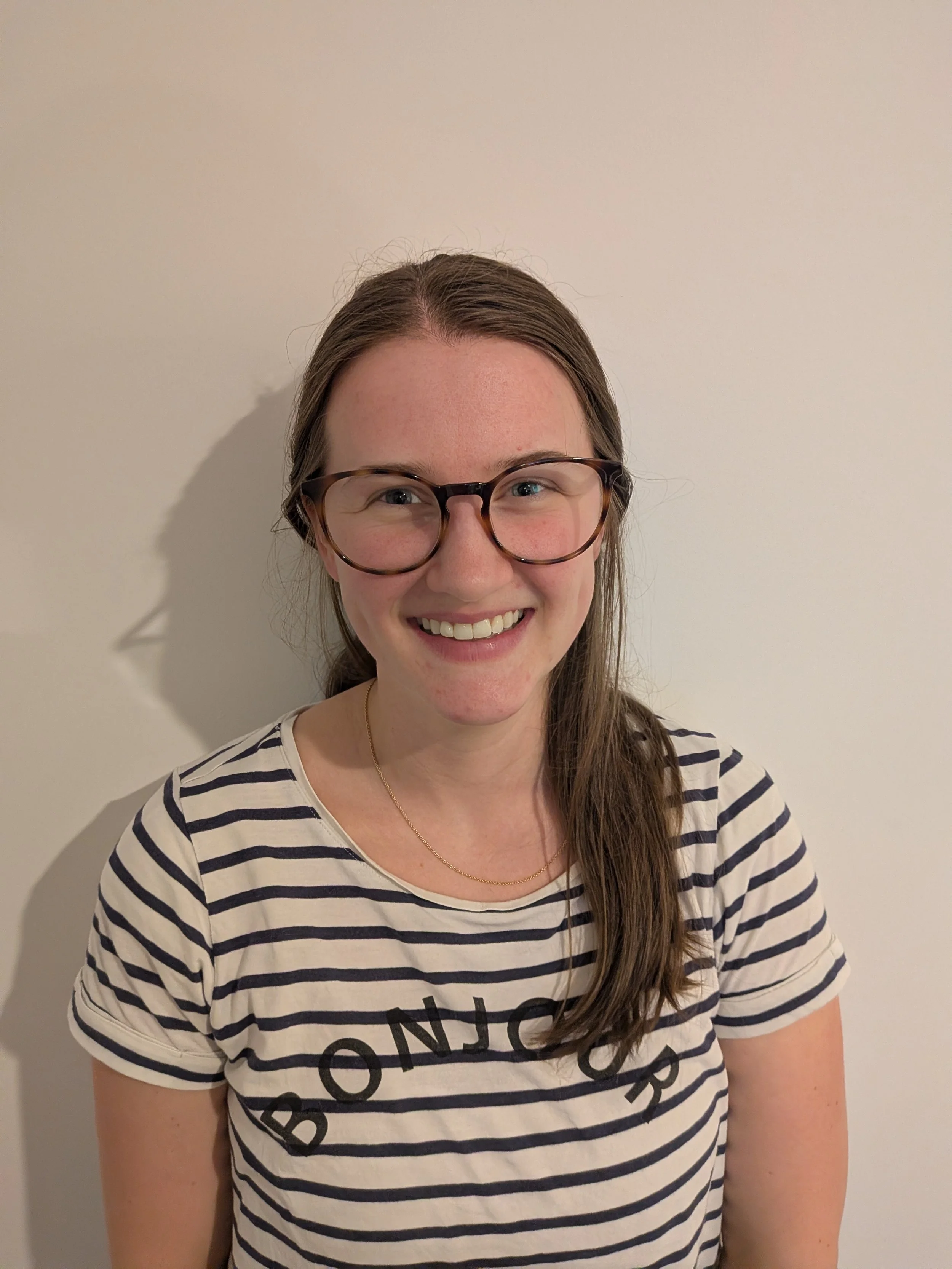 A young woman with long brown hair, wearing glasses and a striped t-shirt, smiling against a plain white wall background.