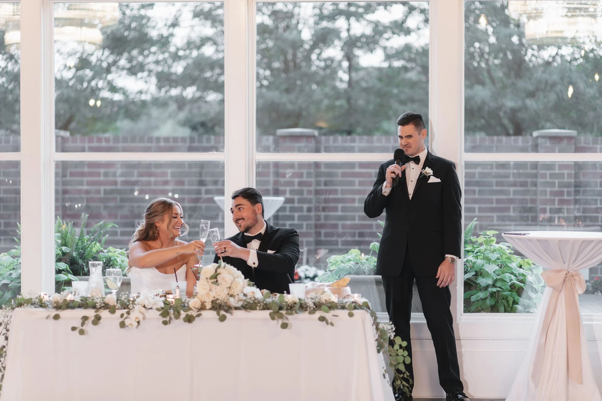 Bride and groom seated at sweetheart table cheersing glasses while best man stands next to them giving speech