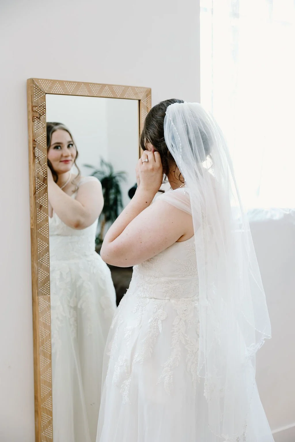 Bride looking in mirror with dress and veil on and putting on earrings while getting ready
