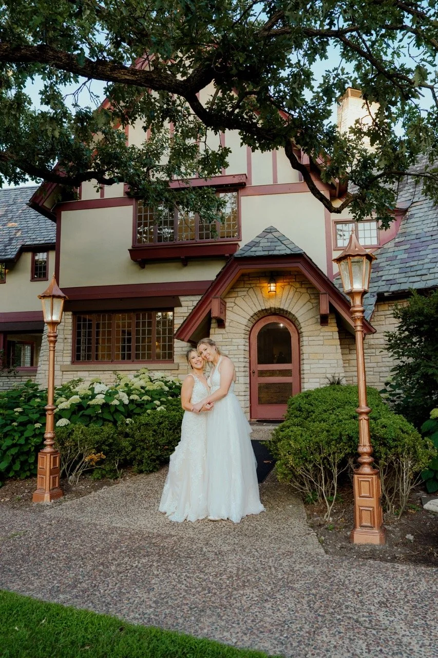 Two brides holding each other smiling with estate house in background