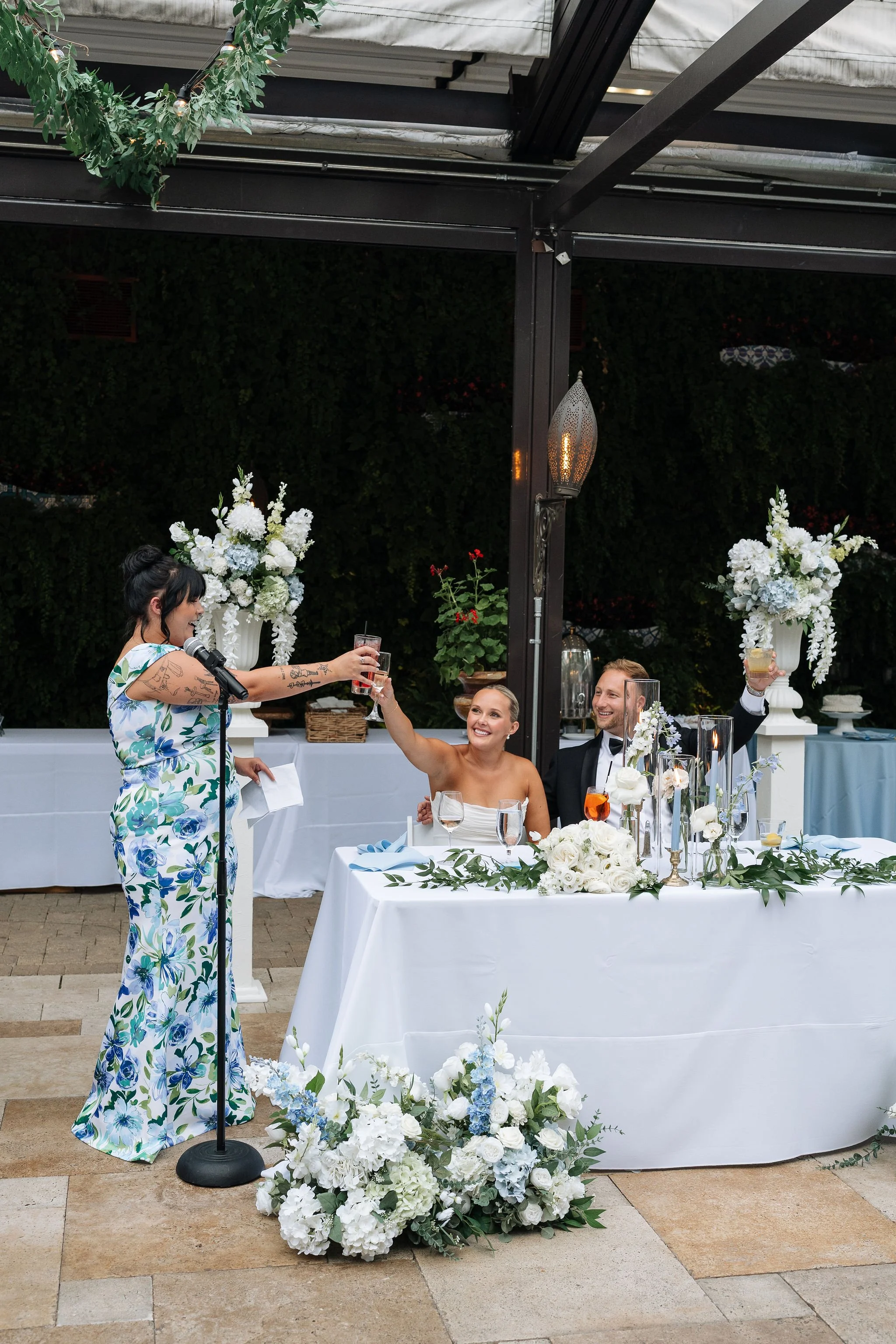 Bride and groom sitting at sweetheart table smiling and cheersing their glass with standing maid of honor after speech