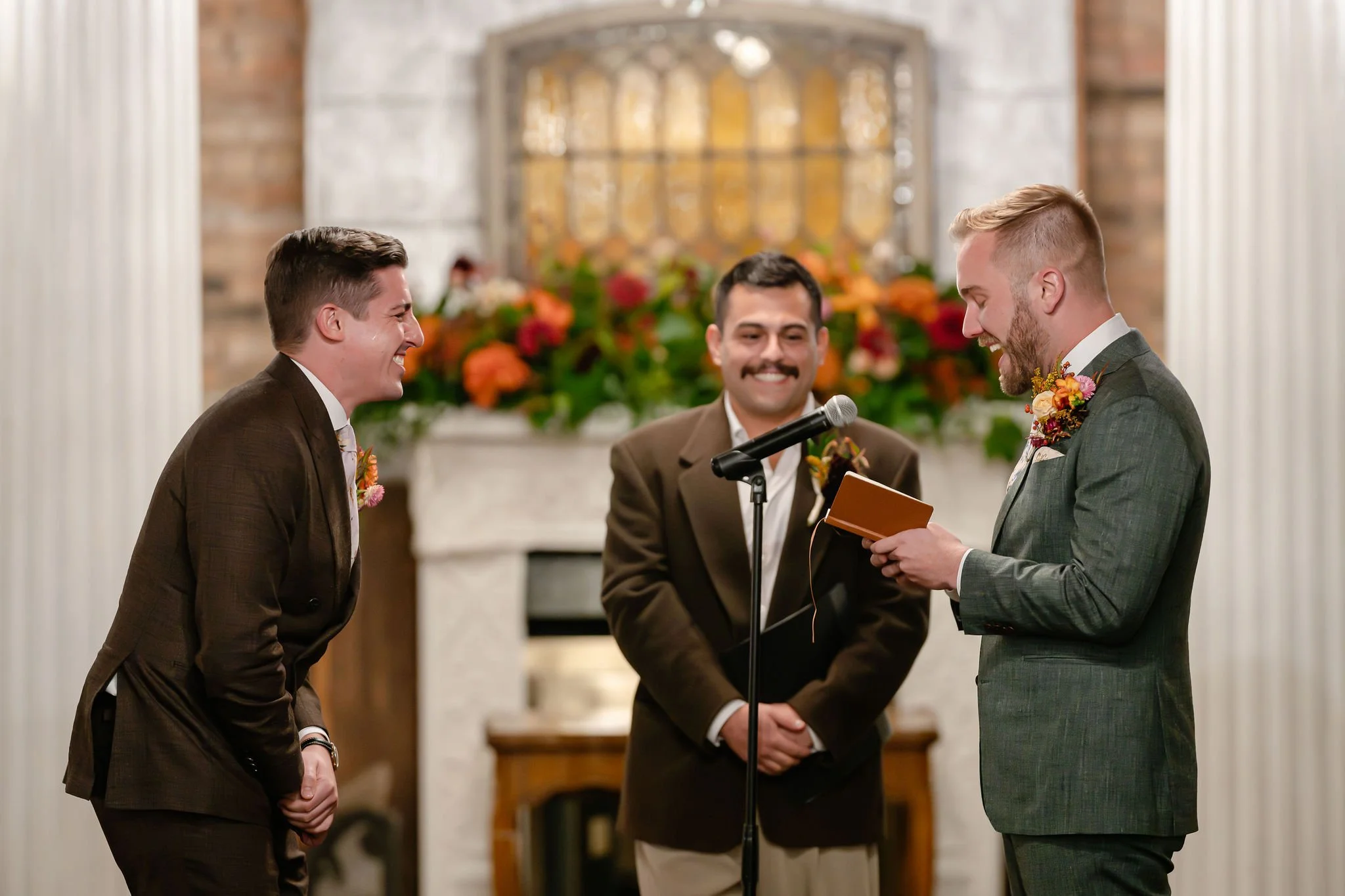 Two grooms at wedding ceremony with one in green reading vows and one in brown suit bent over laughing