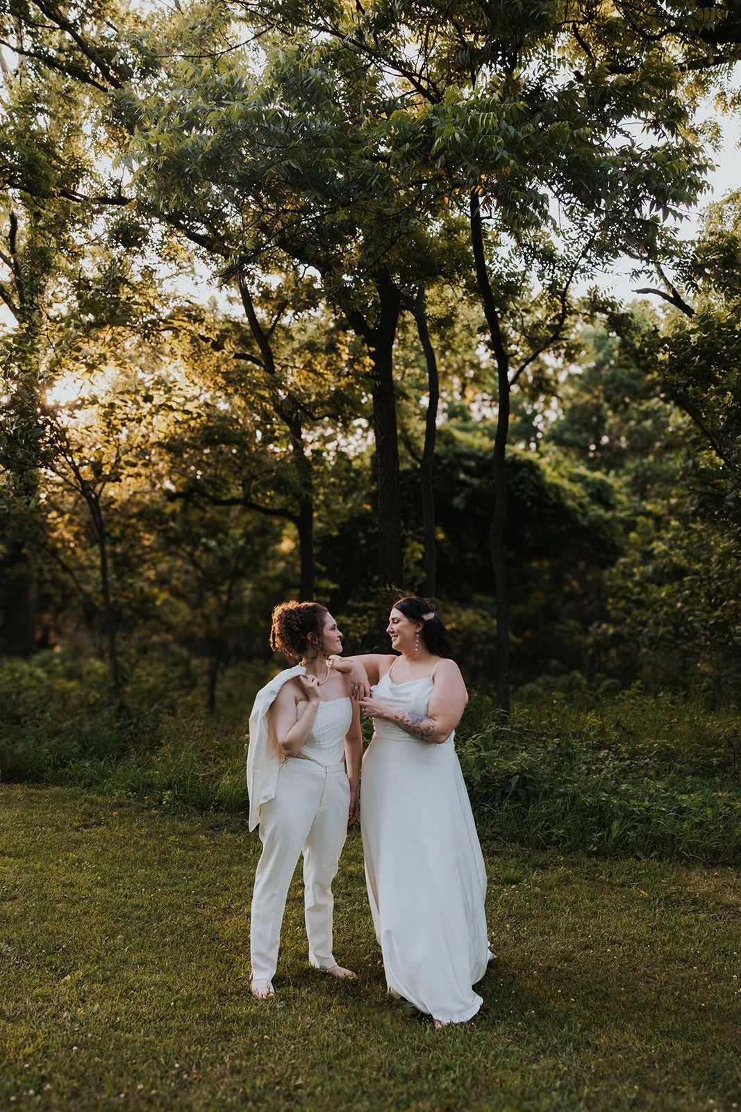 Two brides leaning against each other smiling with bride holding suit jacket over her shoulder
