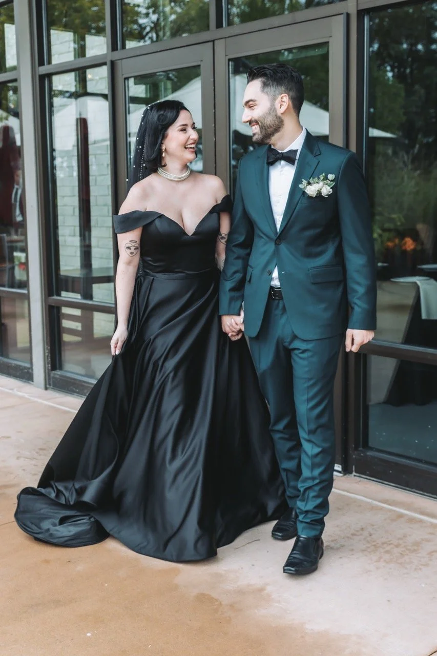 A bride in black wedding dress and groom in green tuxedo holding hands, smiling, indoors near glass doors, with reflections and greenery outside while wedding coordinator works on logistics.