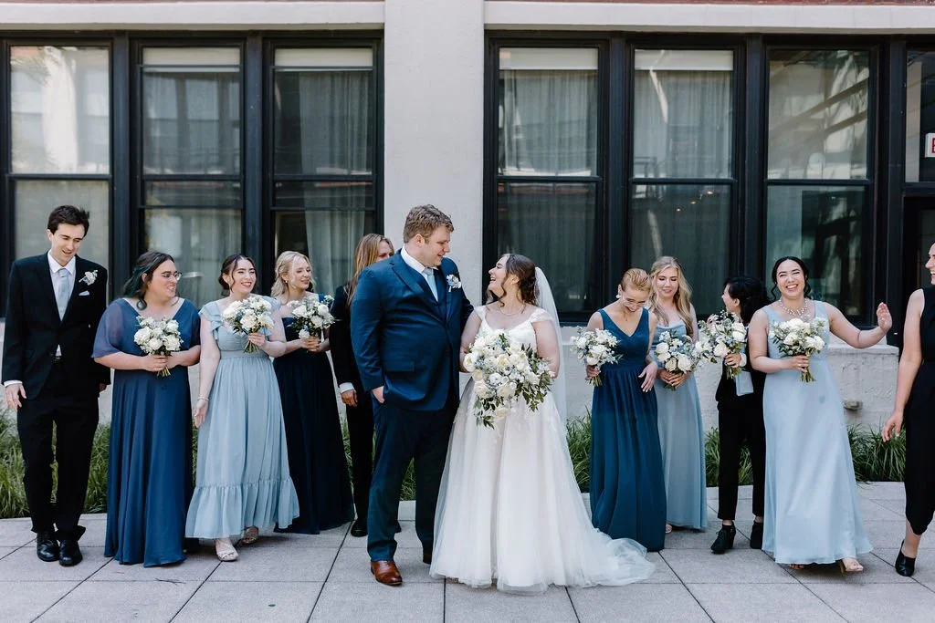 Bride and groom looking at each other while wedding party in dark suits and mismatched blue dresses laugh and look around at each other