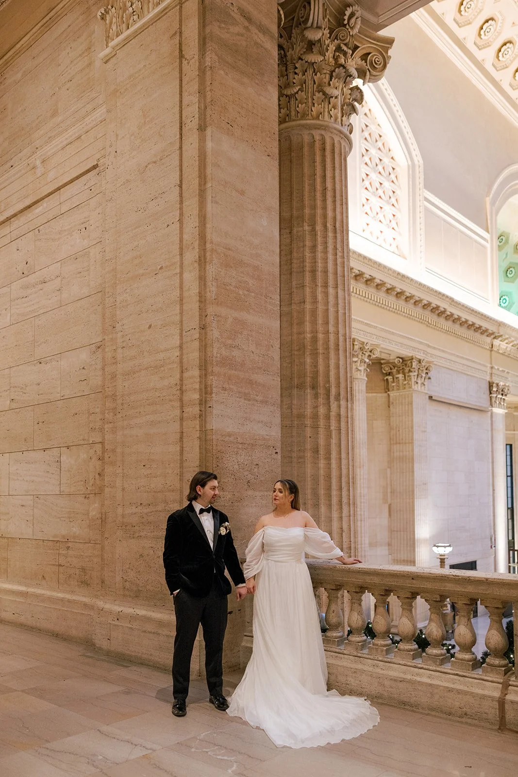 Zoomed out couple holding hands and looking at each other by pillars inside Union Station