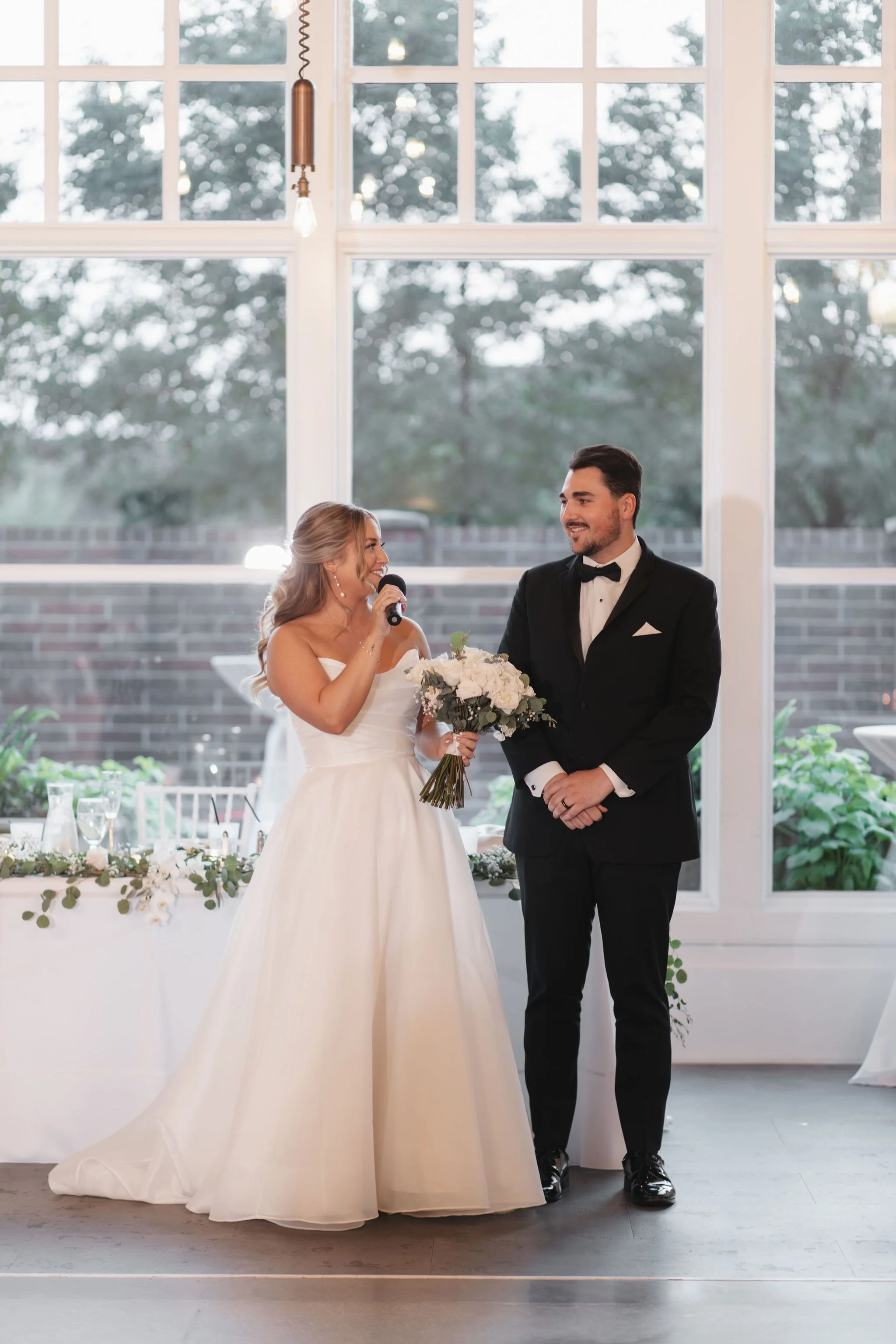Bride holding microphone and bouquet while looking at groom standing in front of a table
