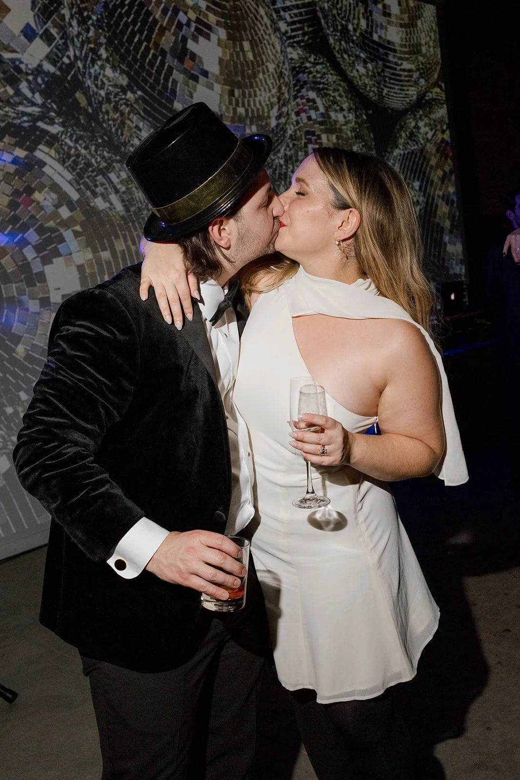 Bride in short reception dress kissing groom with NYE hat holding drinks