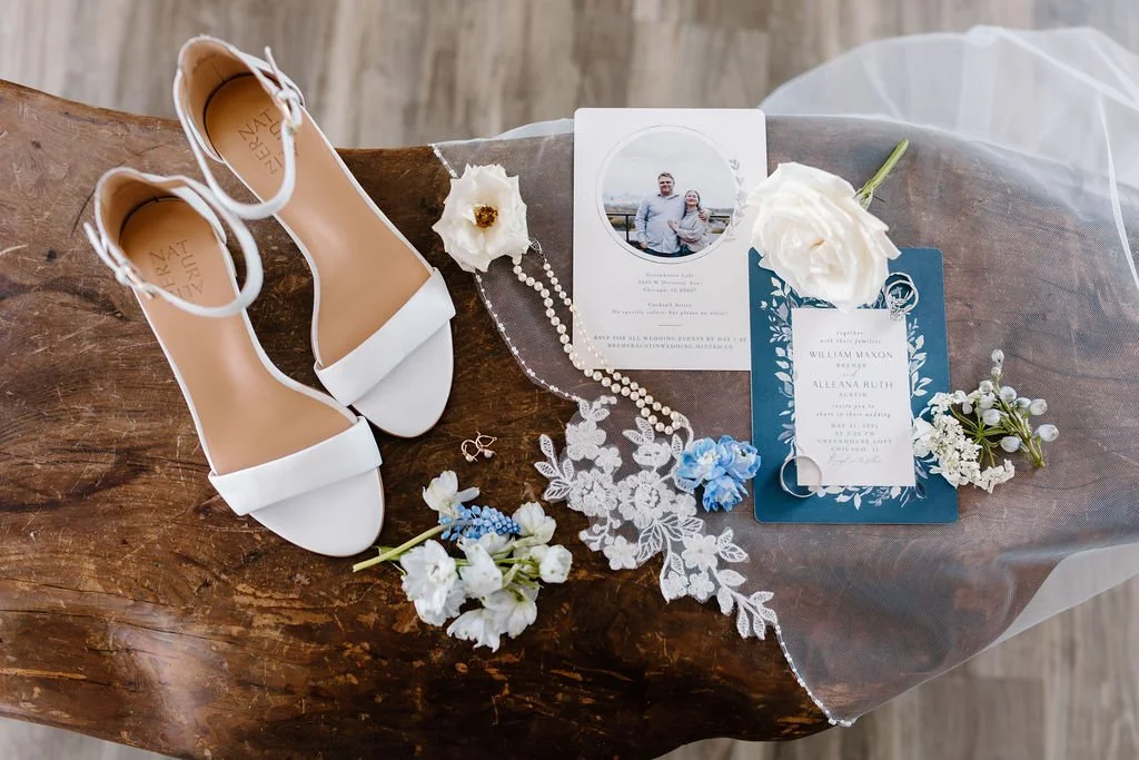 Wedding invitation on veil with shoes, jewelry, and flowers on top of wood table