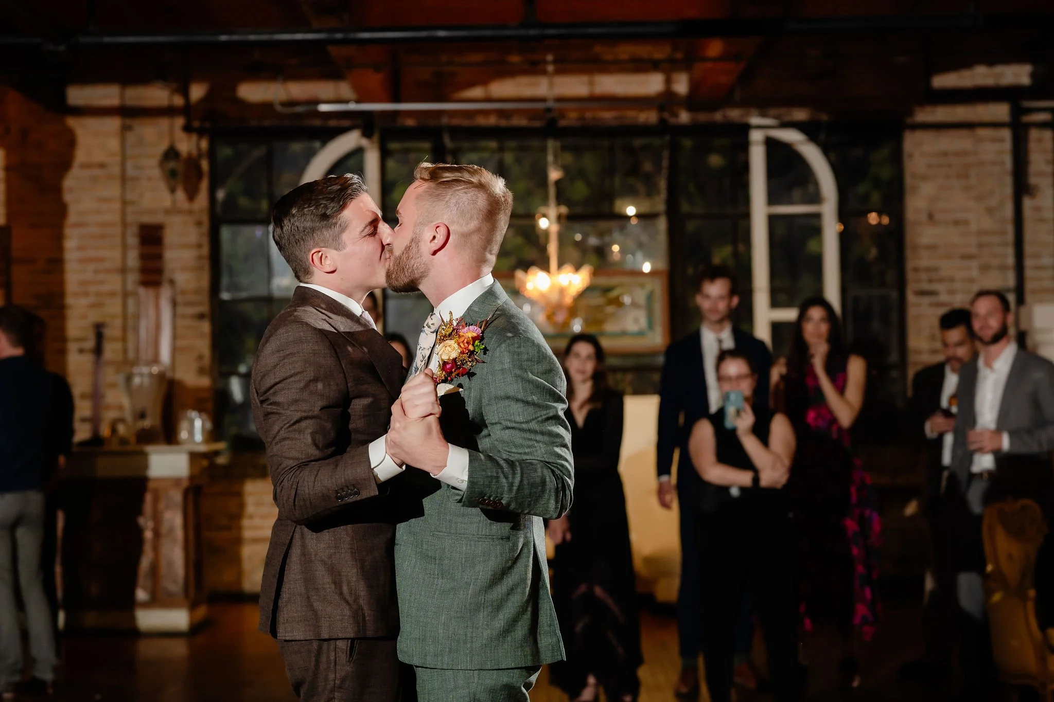 Two grooms kissing on dance floor with family and friends in background