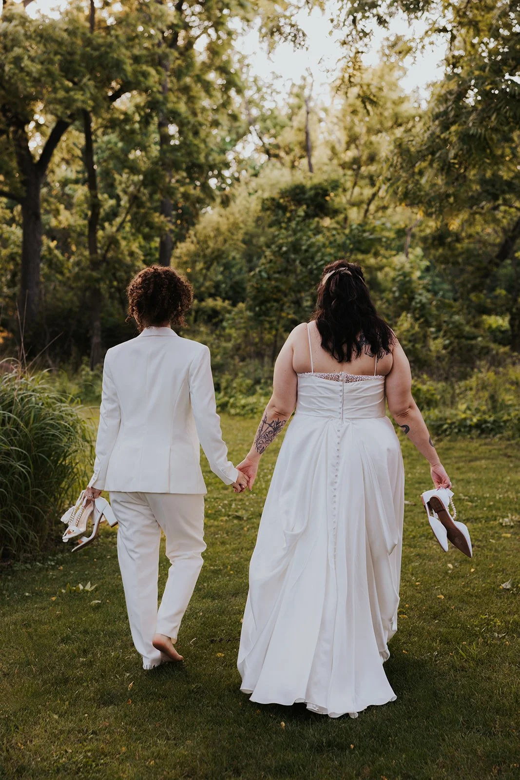 Two brides walking away from camera holding hands and their shoes for sunset photos