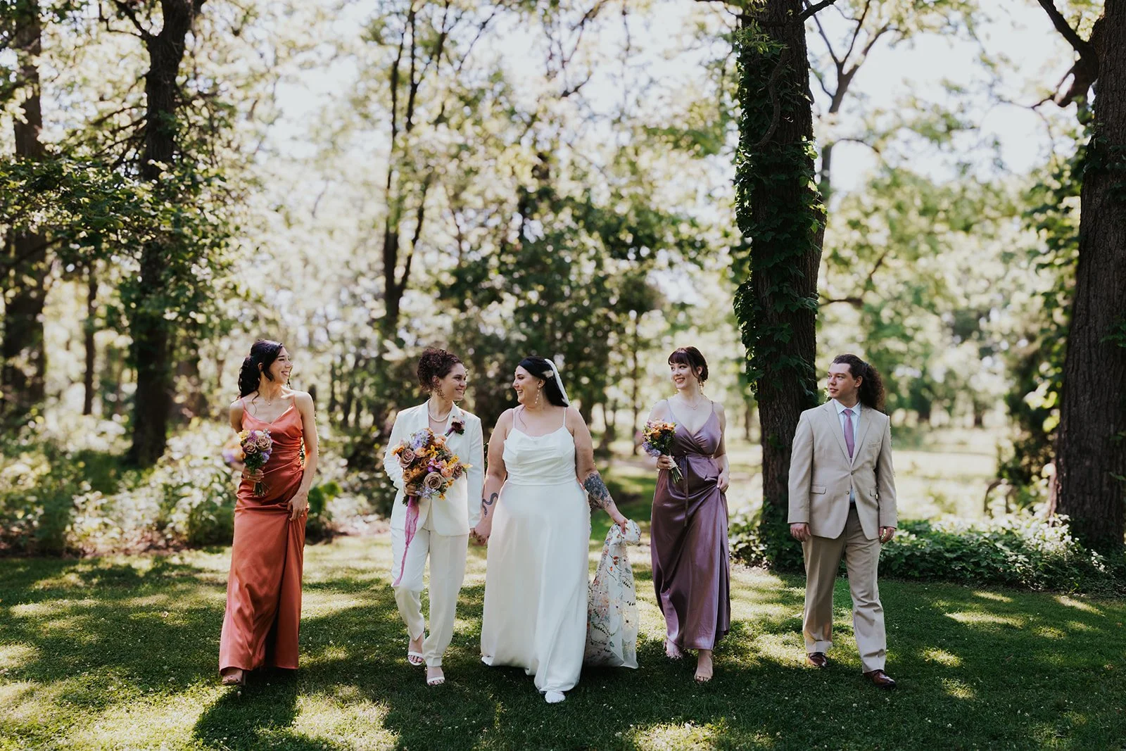 Two brides holding each other while walking with three wedding party members in dresses and suits