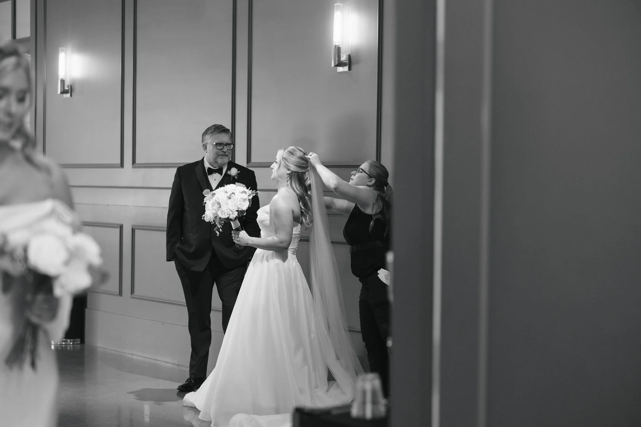 Black and white photo of wedding coordinator putting on veil for bride with father standing watching