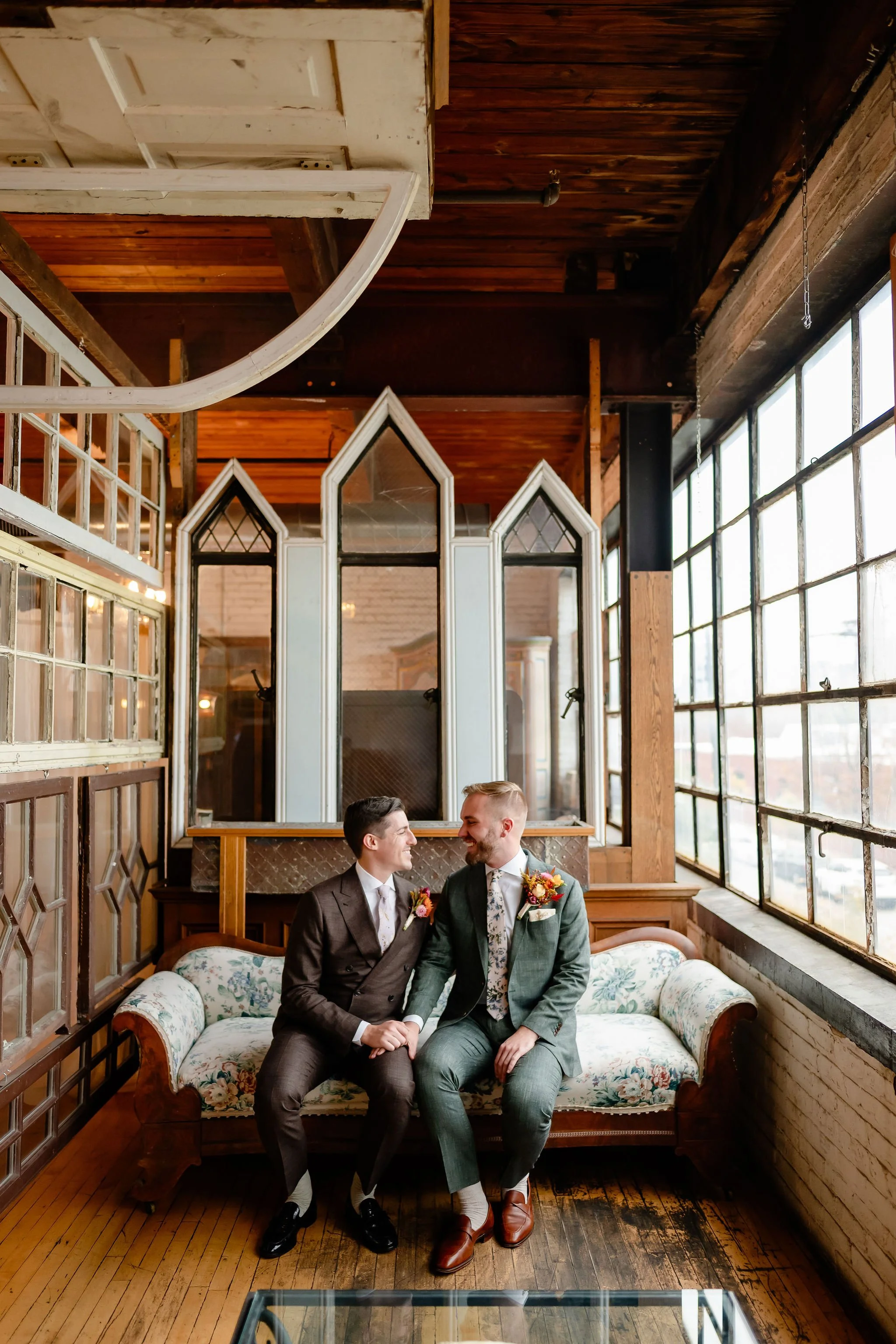 Two grooms sitting on antique couch looking at each other and smiling