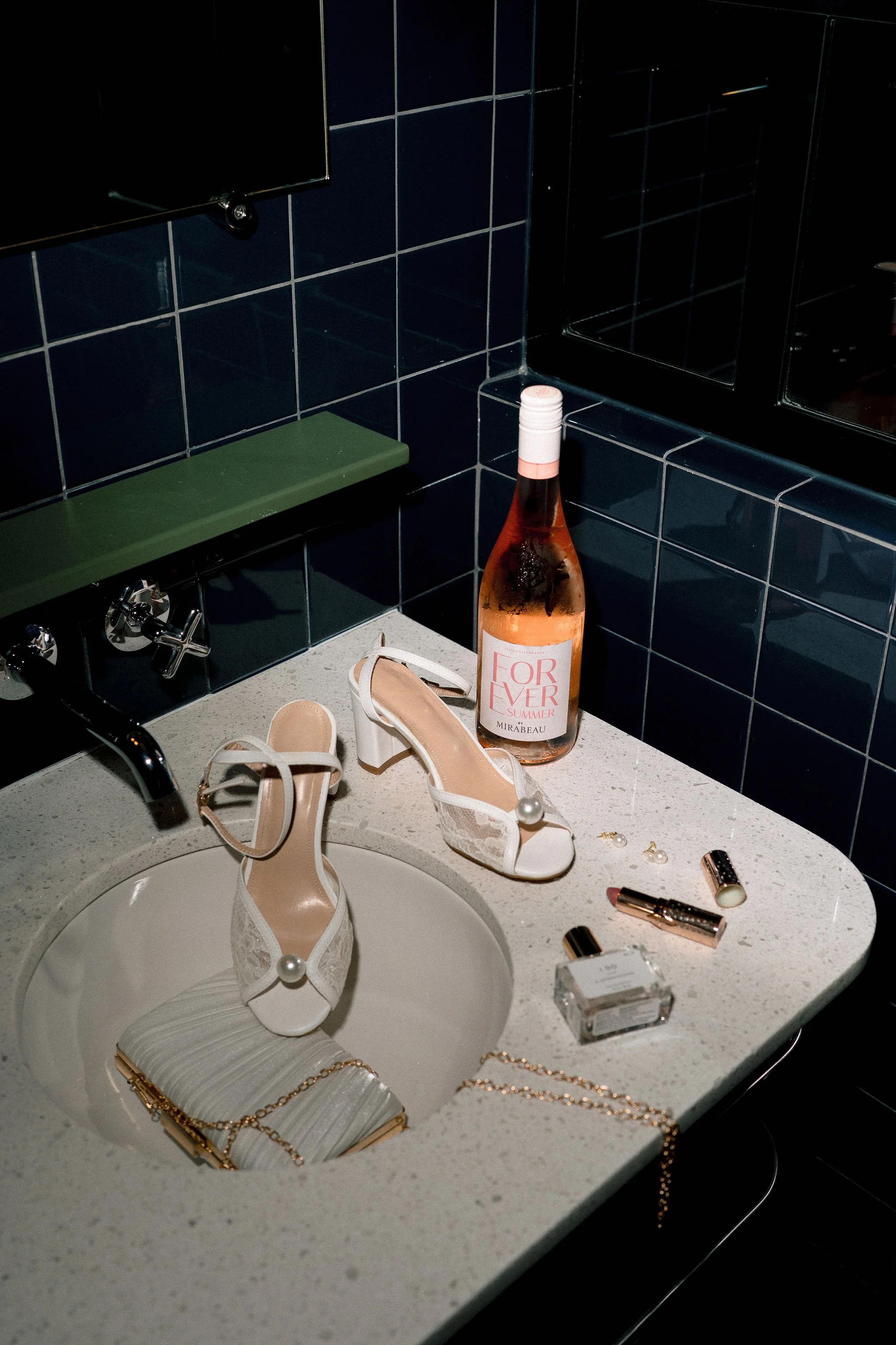 wine bottle, shoes, purse, makeup, and perfume all set in a bathroom sink flash photo editorial style