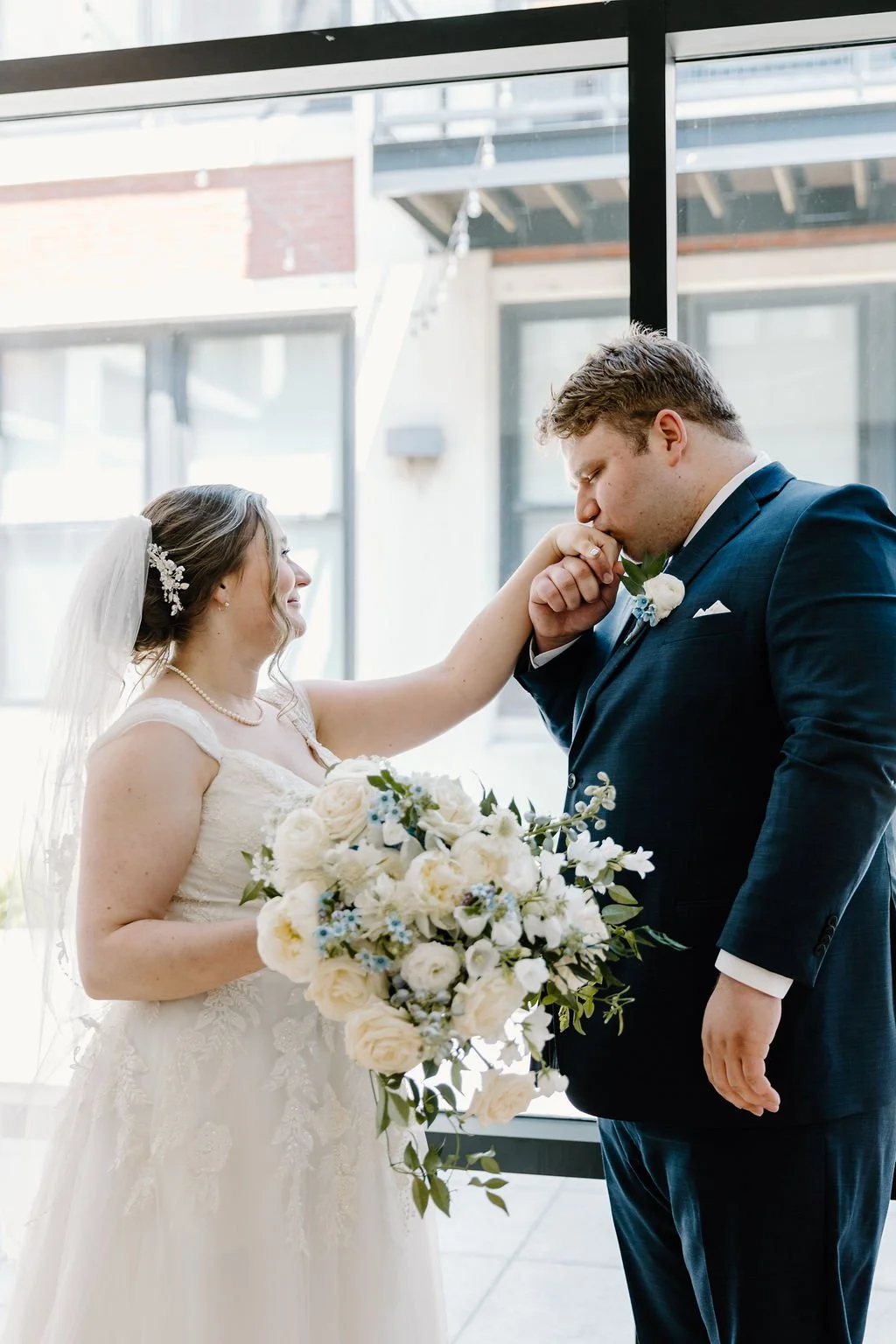 Groom kissing bride's hand while bride smiles holding wedding bouquet