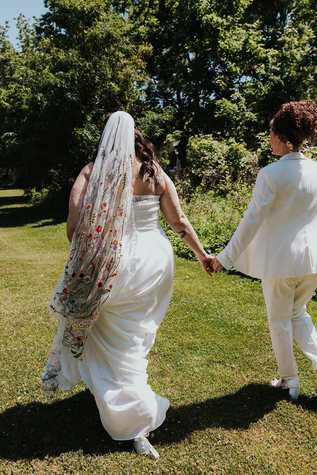 Photo from behind two brides holding hands and bride in dress is holding veil and train