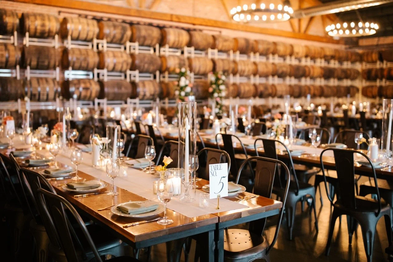 Warm wedding reception room with tables, chairs, candles, and barrels lining background walls