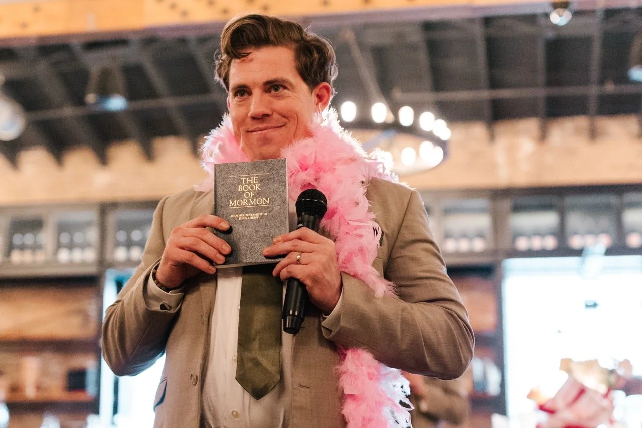 Groomsman in pink scarf holding Book of Mormon and microphone with a grin for wedding speech