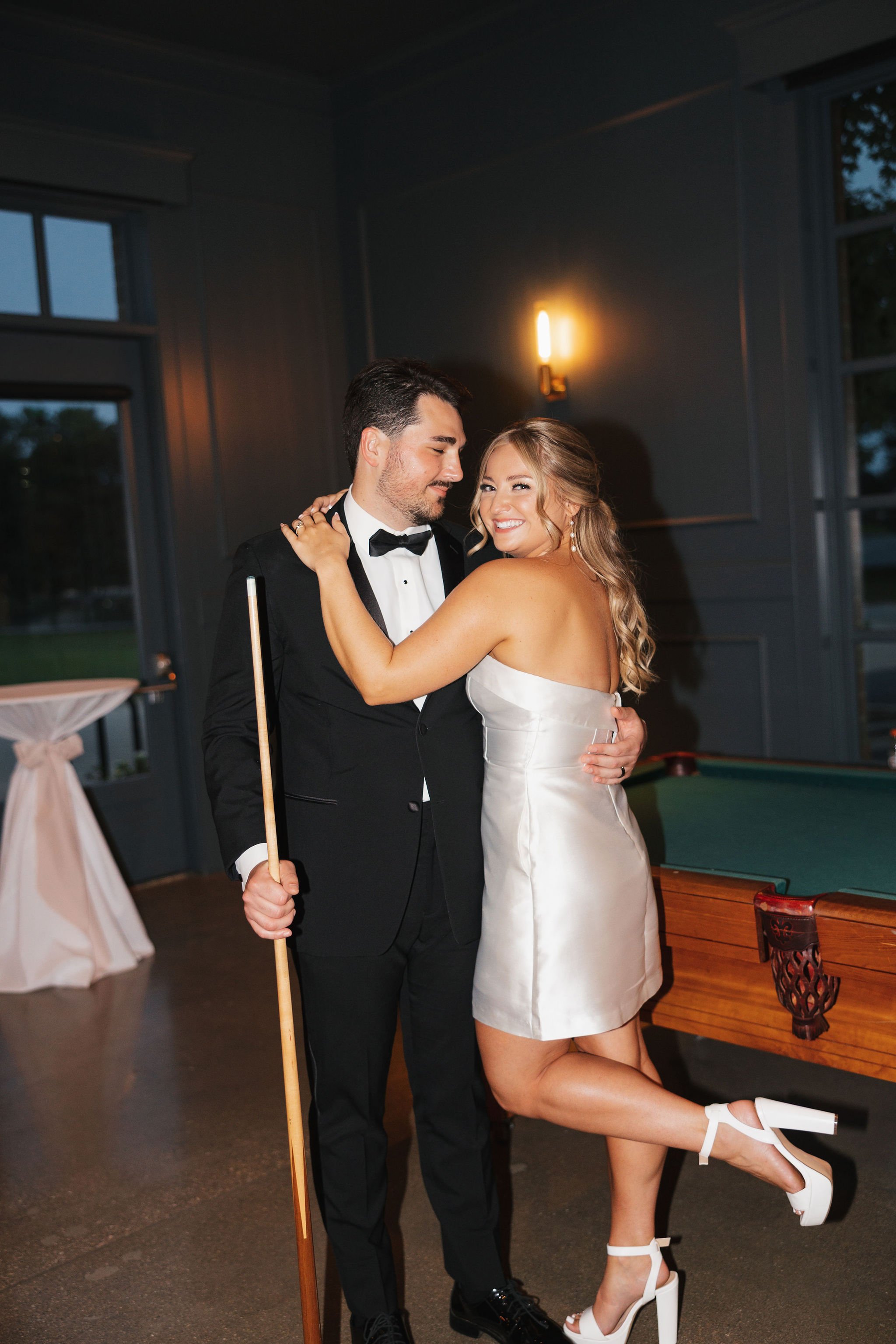 bride leaning against groom while kicking her leg up flash photos in Billiard's room by pool table