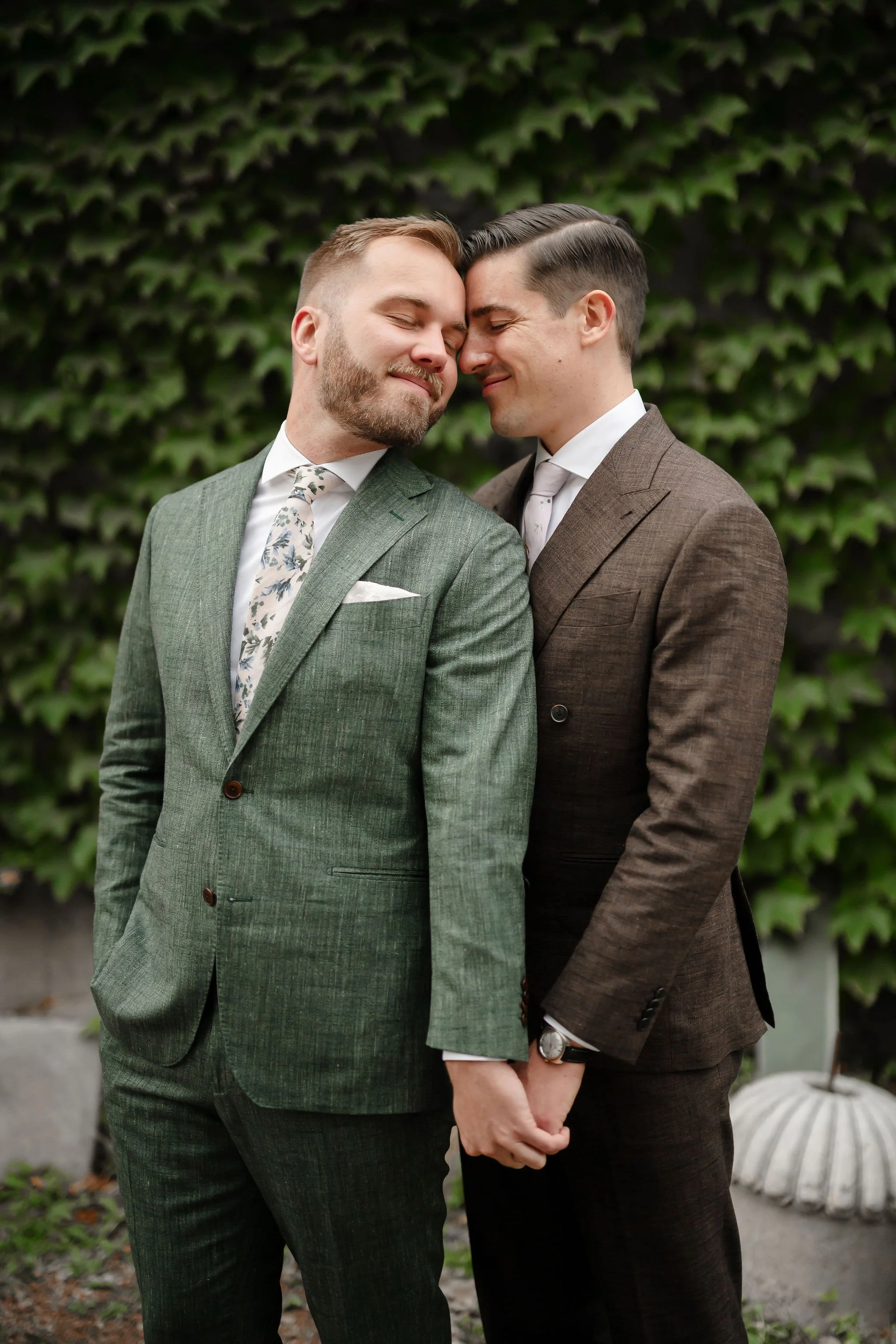 two grooms standing next to each other holding hands with foreheads touching and eyes closed and smiling