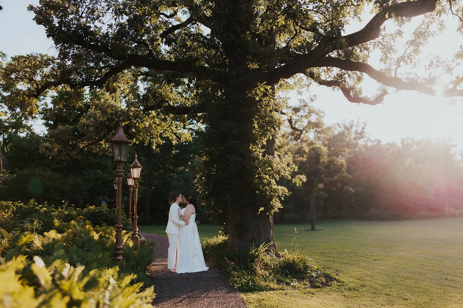 Sunset photos two brides kissing on paved walkway under large oak tree and lamp posts
