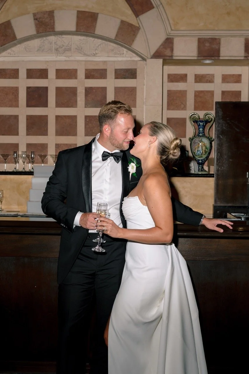 bride and groom leaning against bar in venue smiling at each other almost kissing