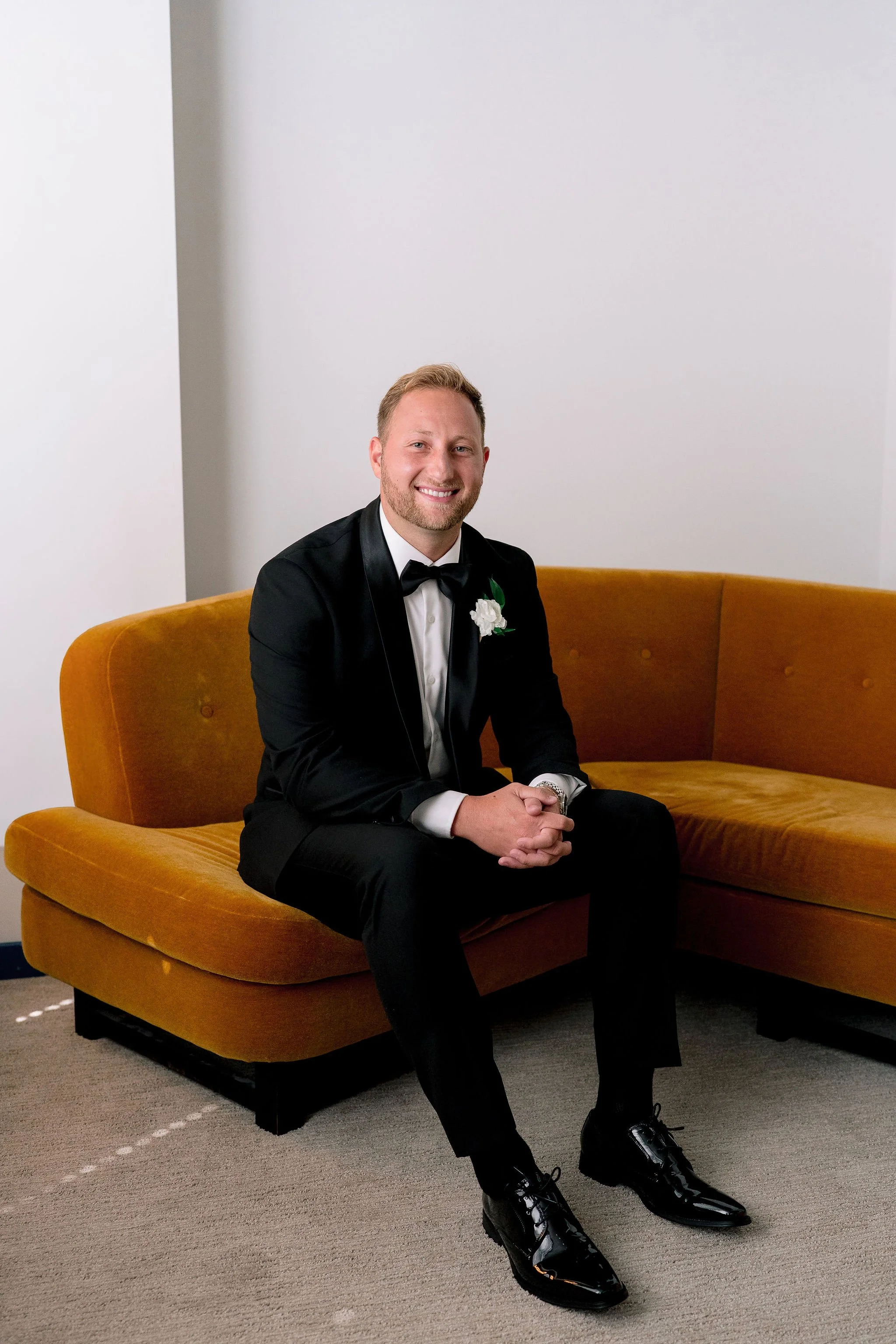 Groom in suit smiling while sitting on an orange couch with a white background