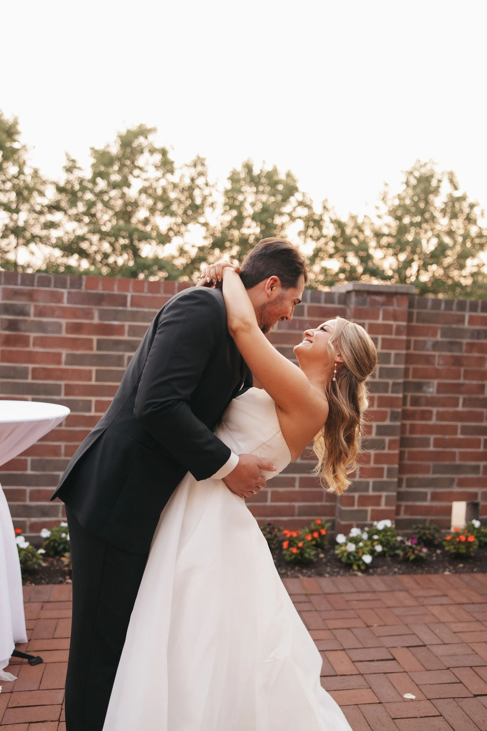 Groom holding and dipping bride while smiling at each other outside with brick floors and wall
