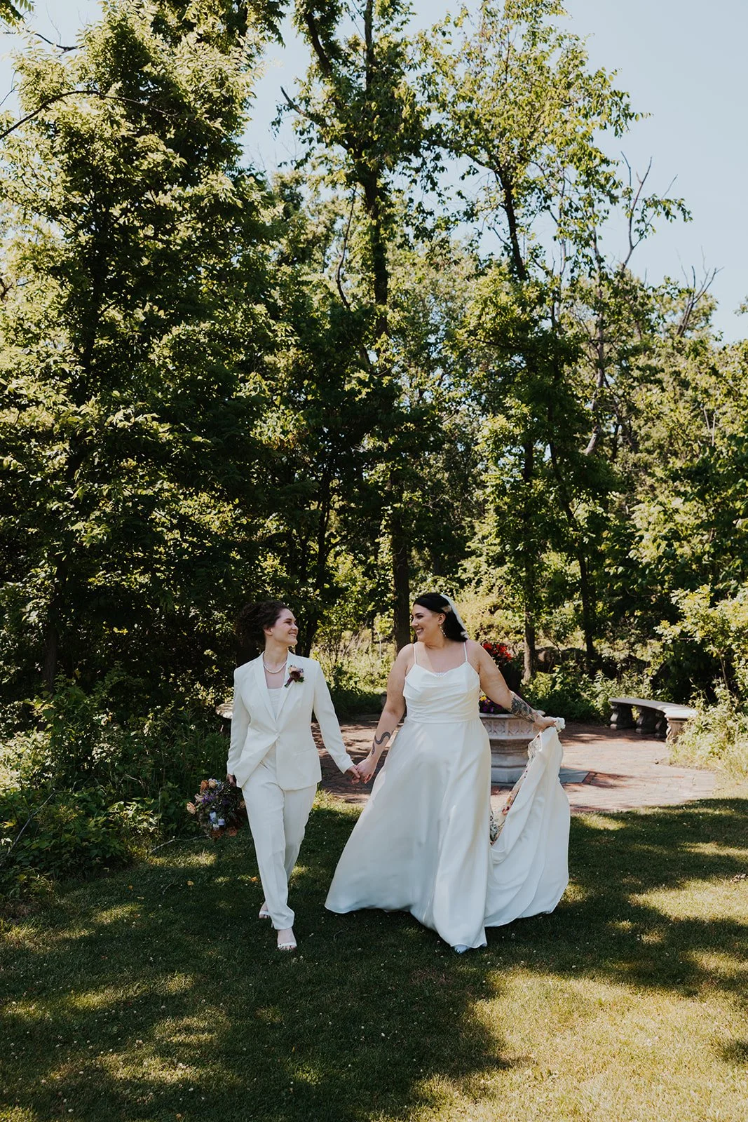 One bride in dress holding train and veil and holding hands with bride in white suit