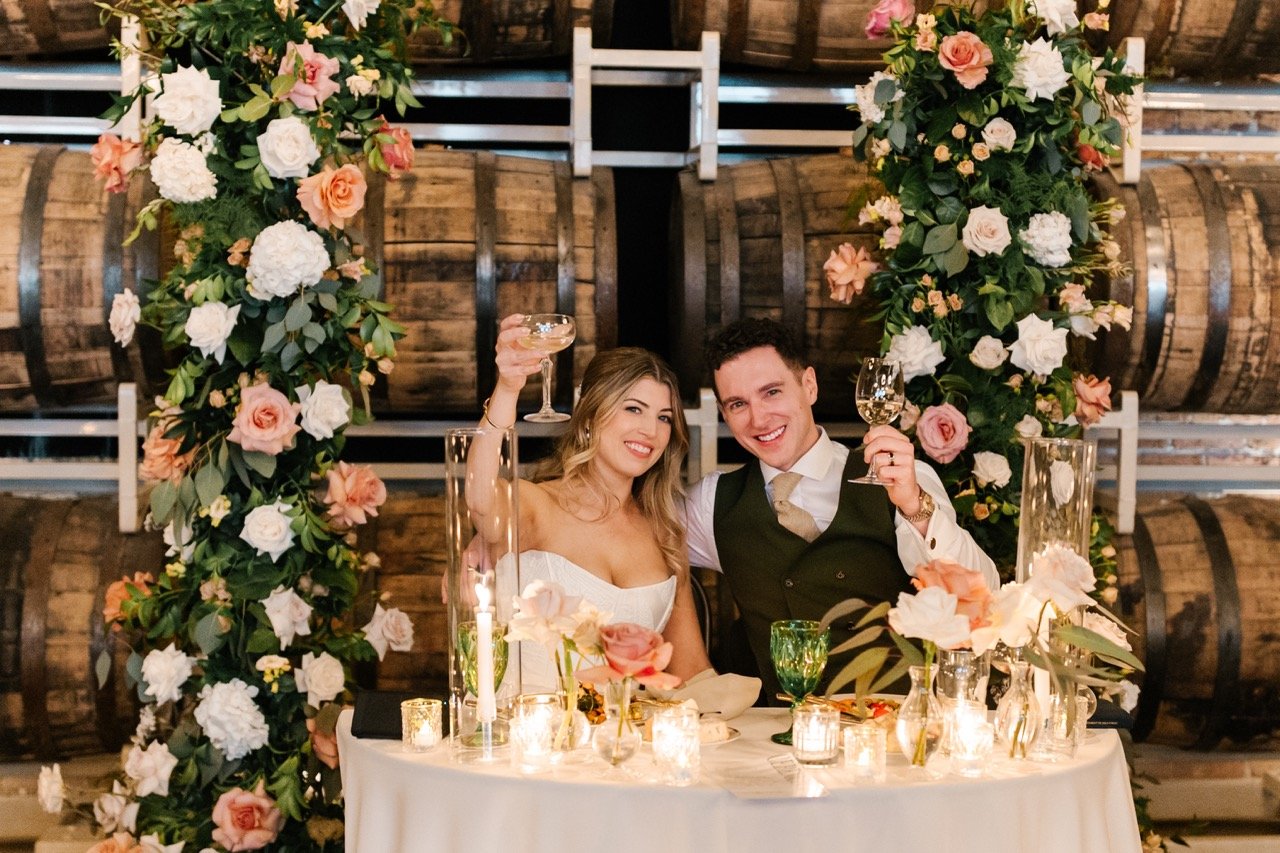 Couple seated at sweetheart table holding up drinks at wedding reception
