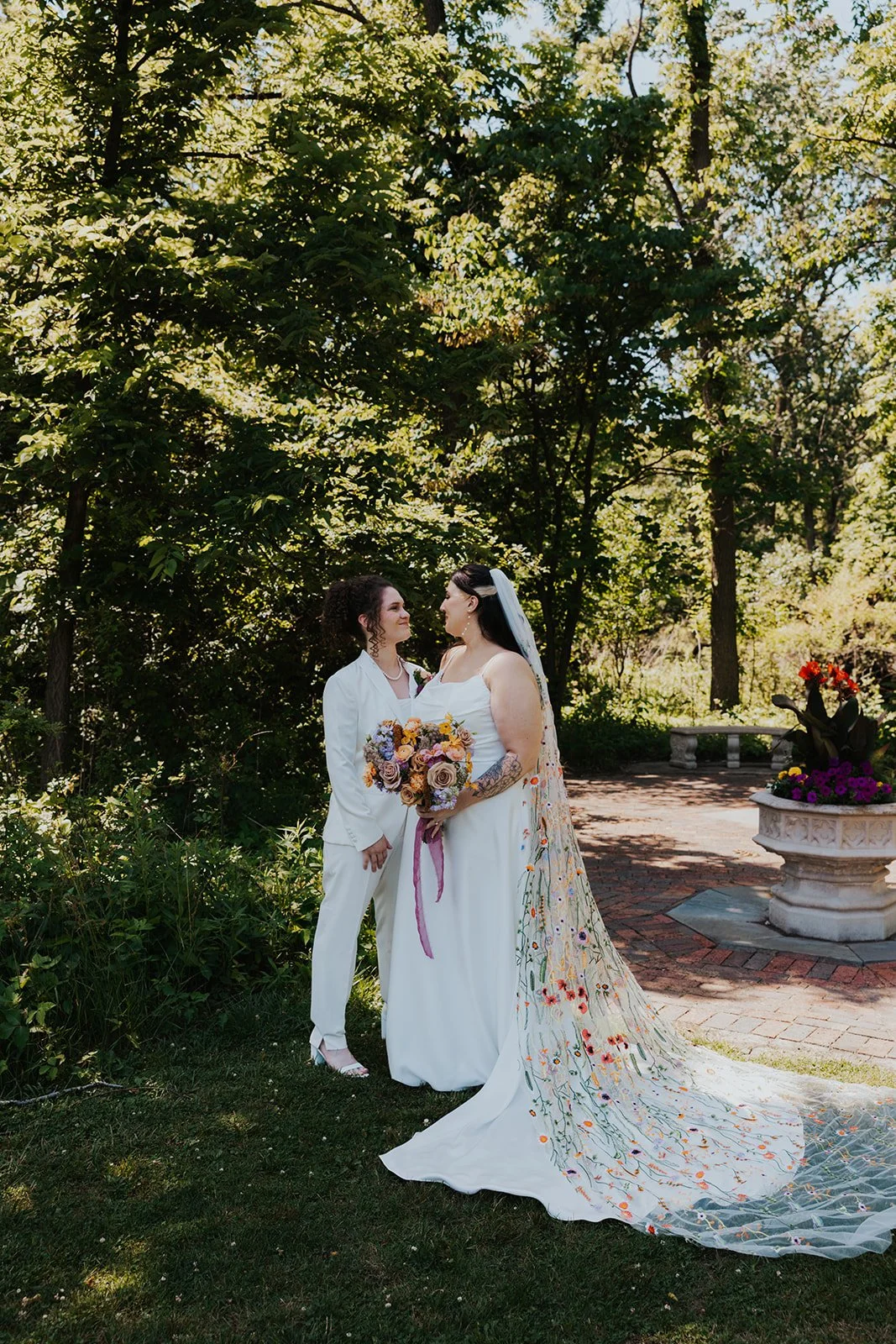 two brides looking at each other and grinning showing off one bouquet and embroidered veil