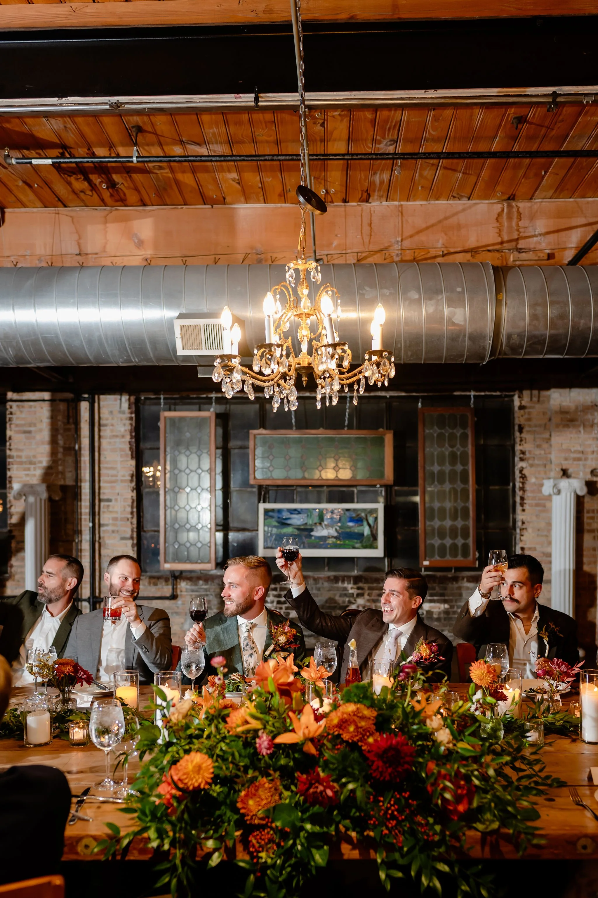 Two grooms and wedding party members at head table cheersing for speeches raising glasses with everyone