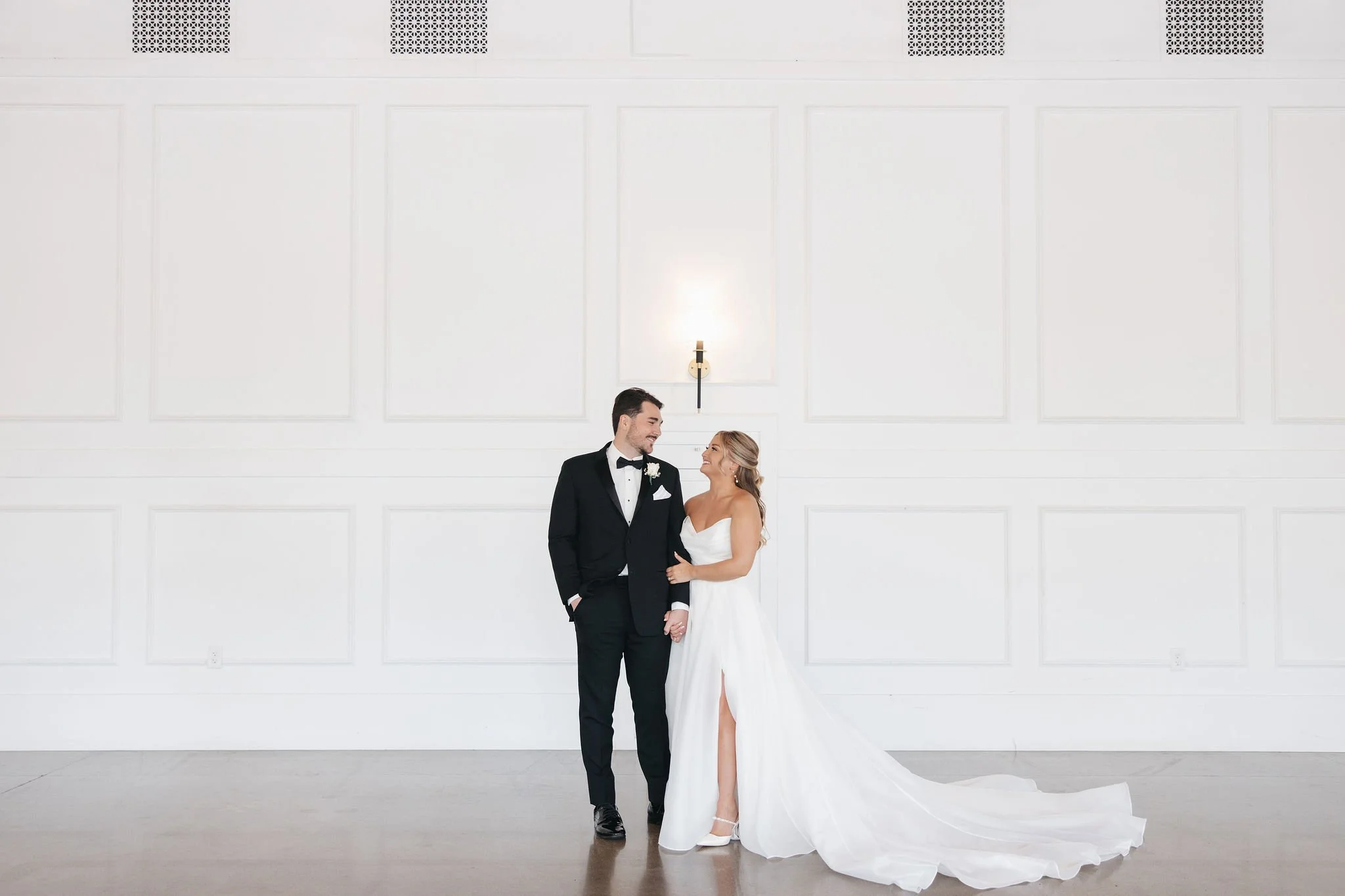Bride and groom holding and smiling at each other against a white wall as a background