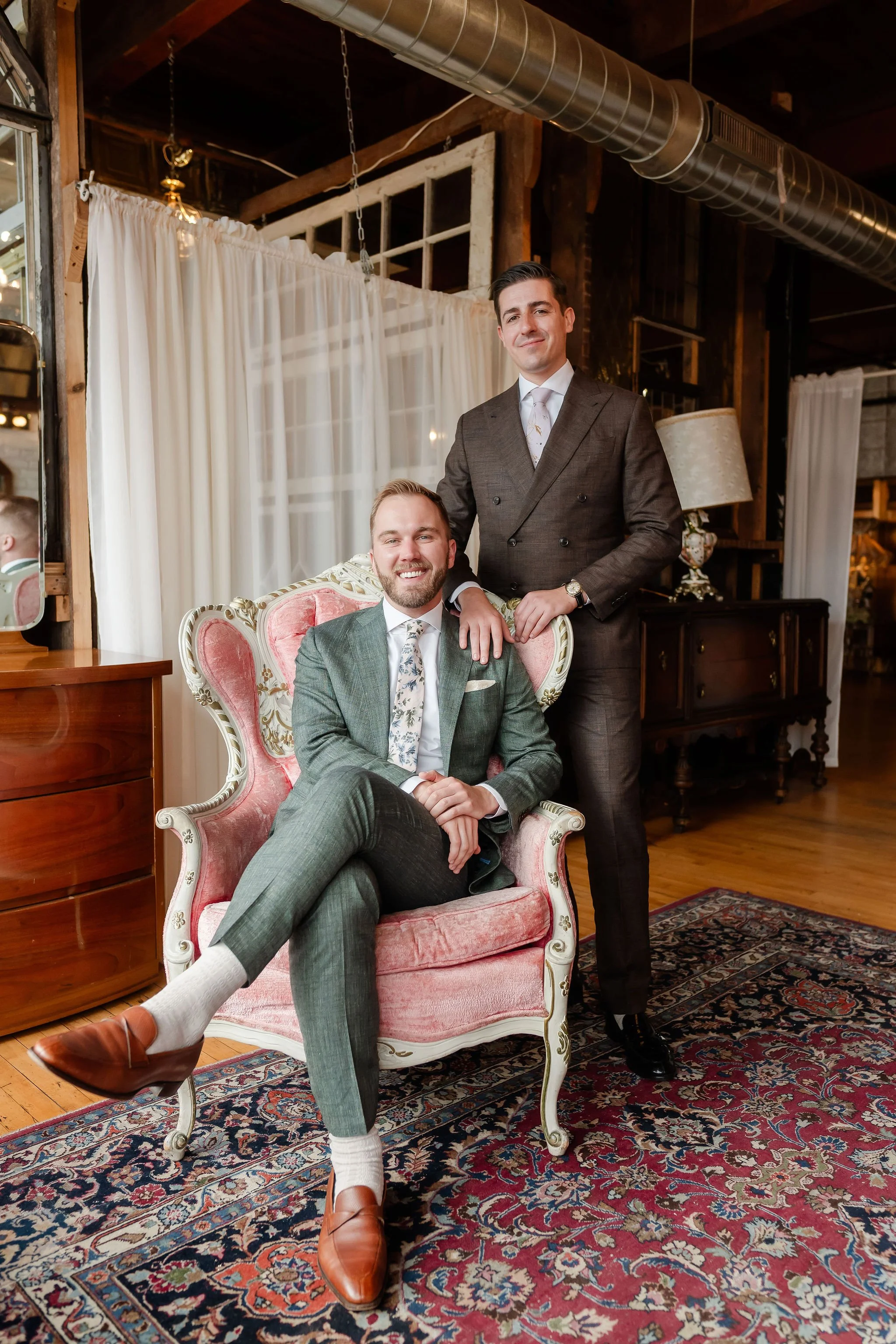 One groom sitting in pink vintage chair with second groom standing with hand on shoulder