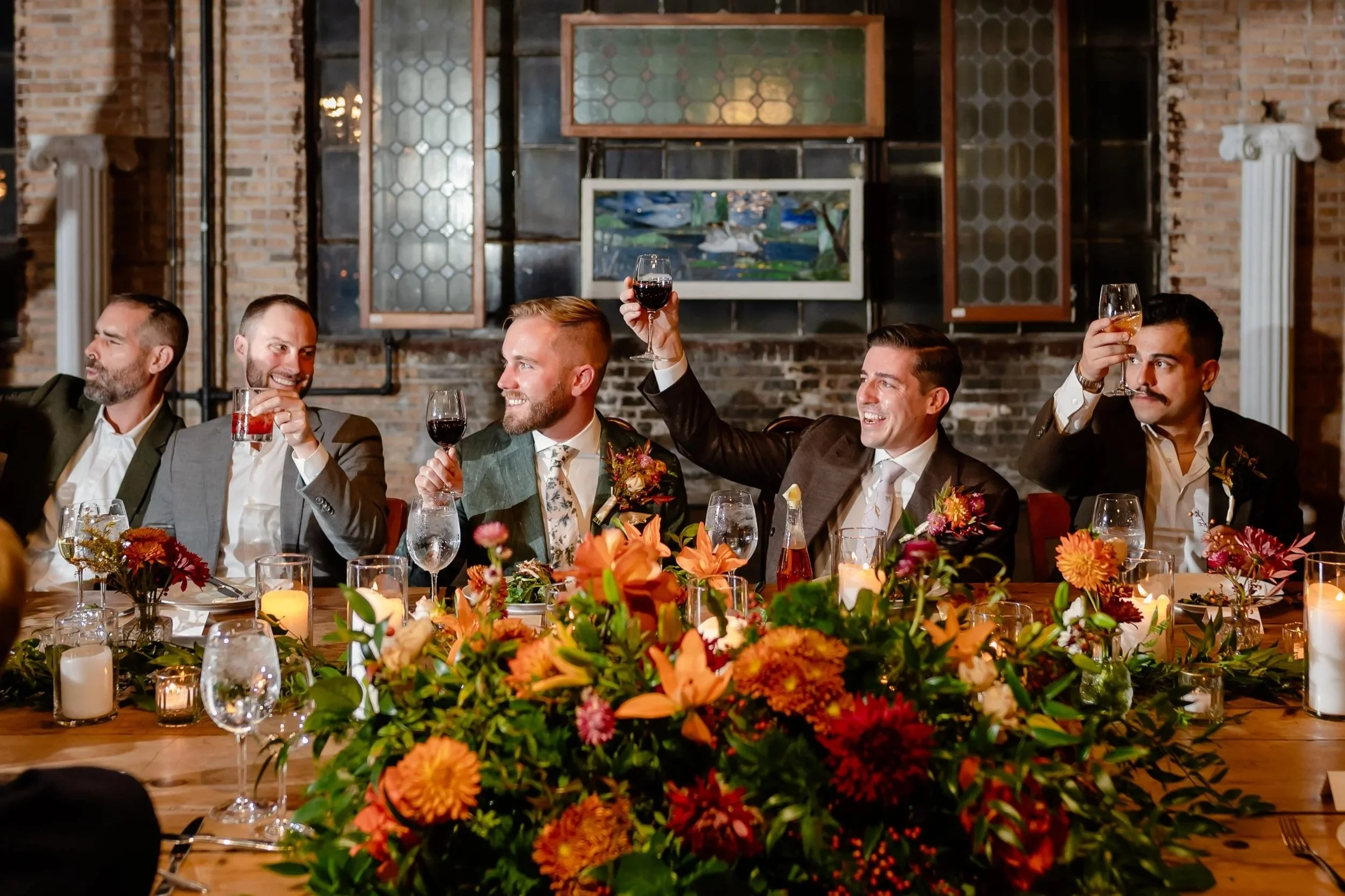 2 grooms sitting with wedding party at reception cheersing their glasses for a toast or speech