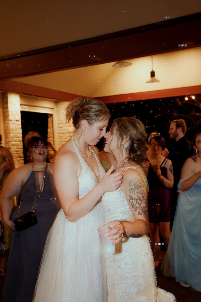 Two brides almost kissing on dance floor with wedding guests dancing