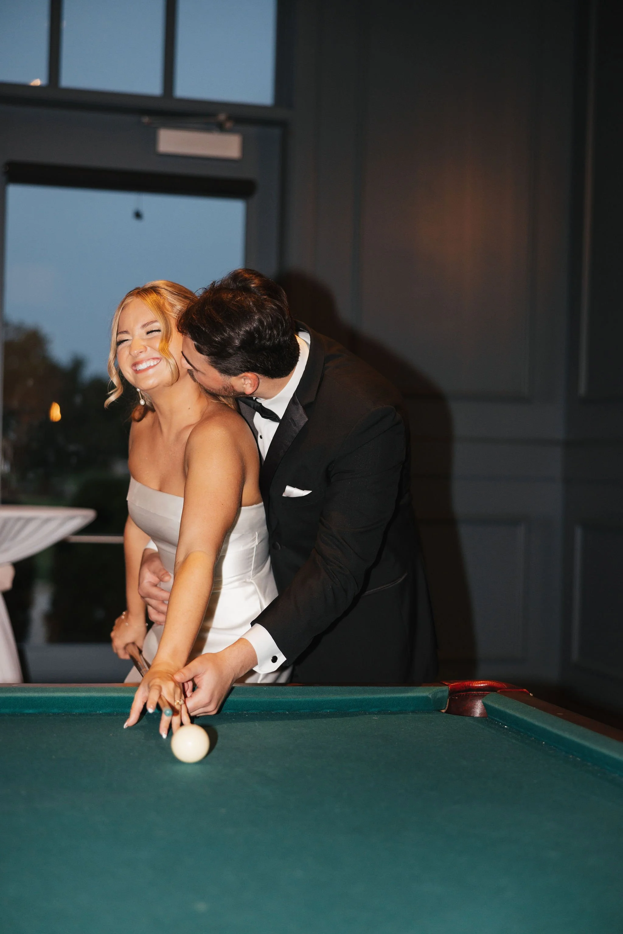 Bride smiling hitting ball at pool table while groom is kissing her