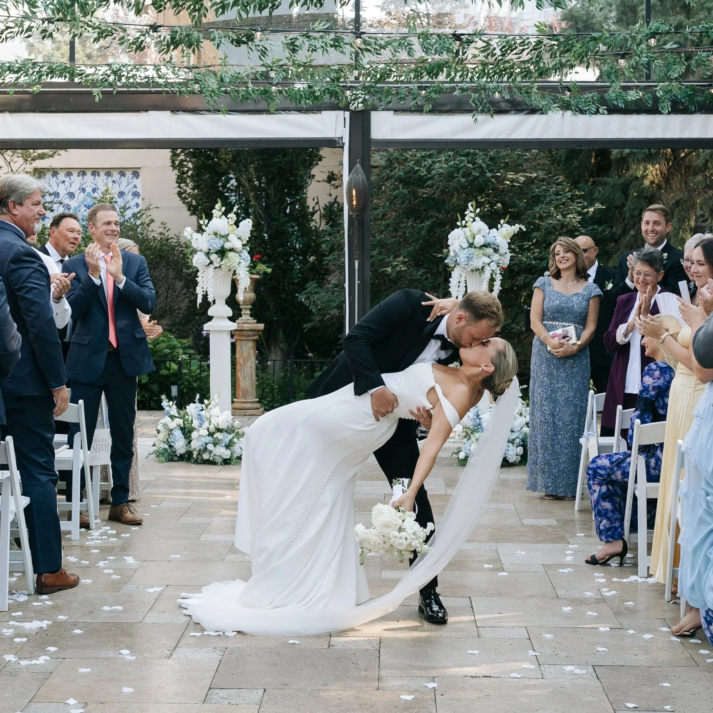 A newlywed couple being helped by a Chicago day-of wedding coordinator sharing a romantic kiss during their wedding ceremony, surrounded by friends and family clapping and smiling, with floral arrangements and outdoor decorations in a garden setting.