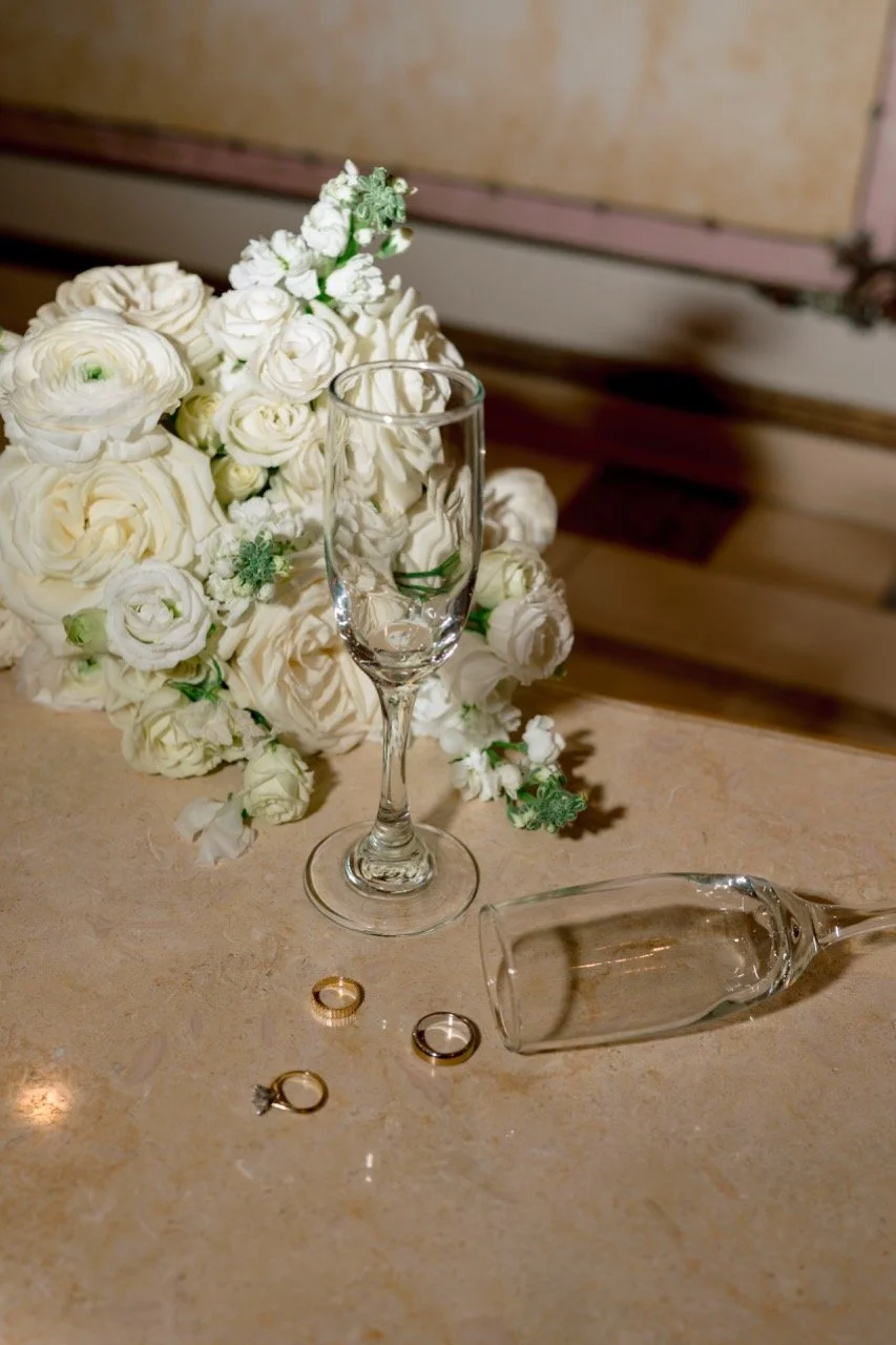 flash photo of details bouquet, two champagne glasses with one tipped over spilling out 3 wedding rings