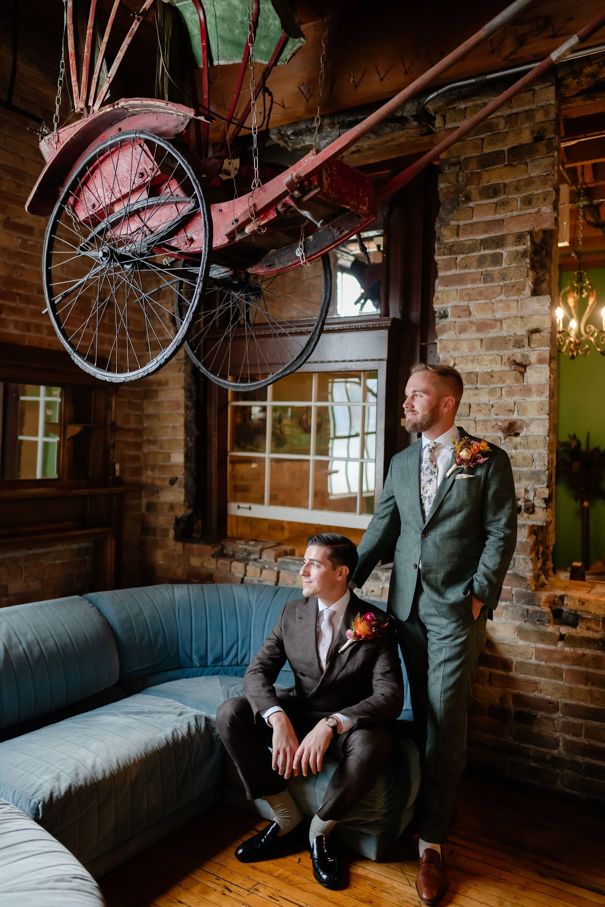 Two grooms, one sitting on antique, long couch and one standing under antique wagon