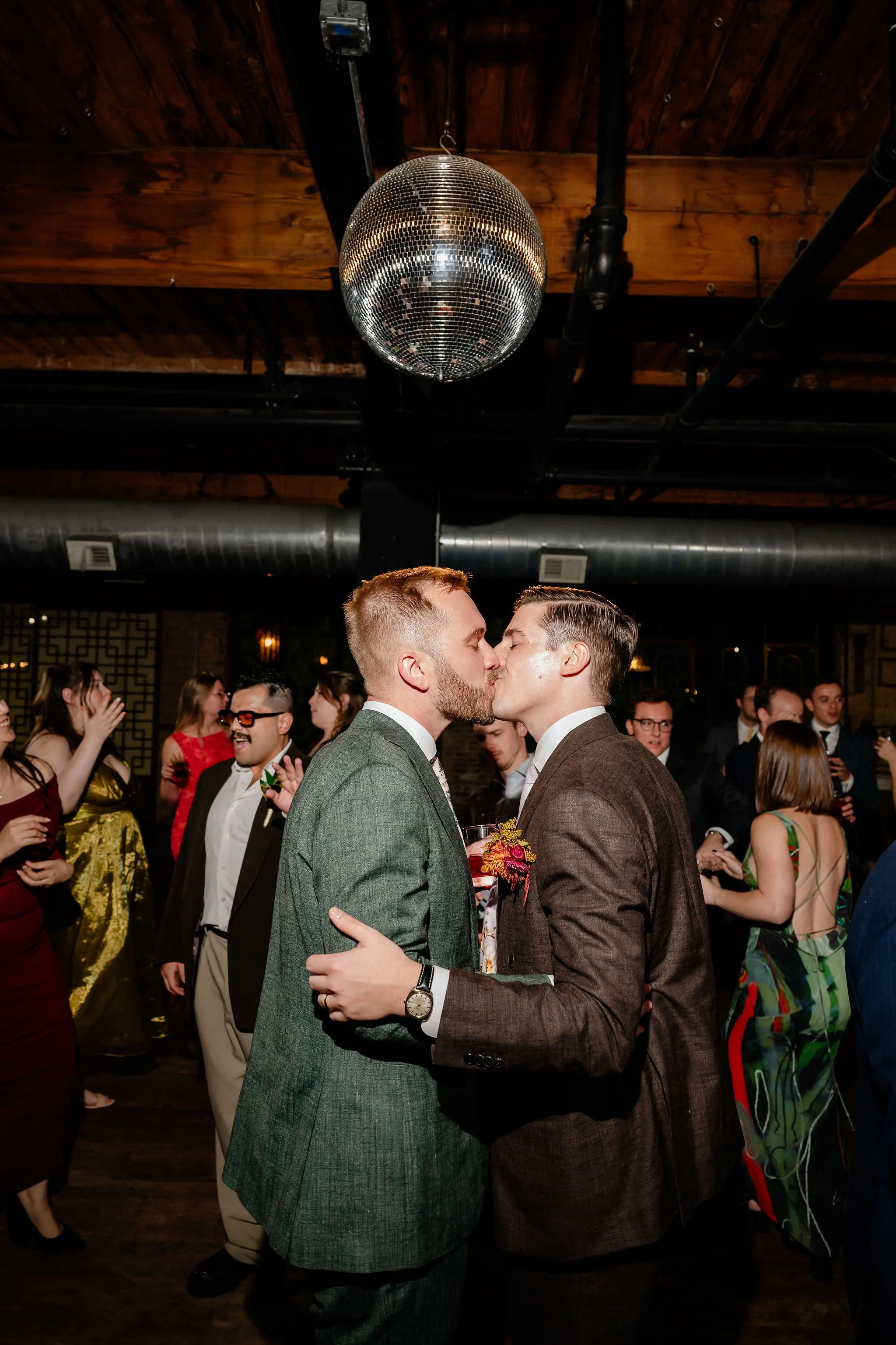 Two grooms kissing on the dance floor with disco ball above wedding guests in background having fun and dancing
