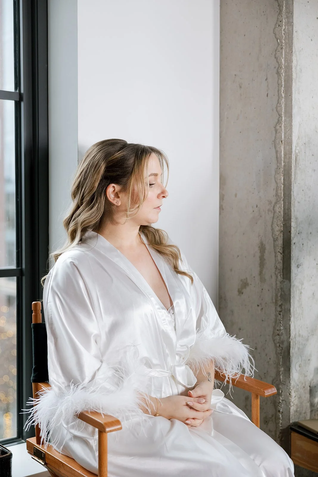 Bride sitting in chair getting ready with white feathers and satin white robe