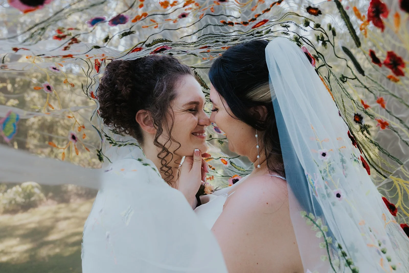 two brides close up smiling and almost kissing underneath embroidered veil